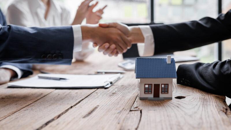 Immobilienmakler und Person geben sich die Hand über einem Hausmodell, Symbol für Baufinanzierung, Erwerb eines Eigenheims.